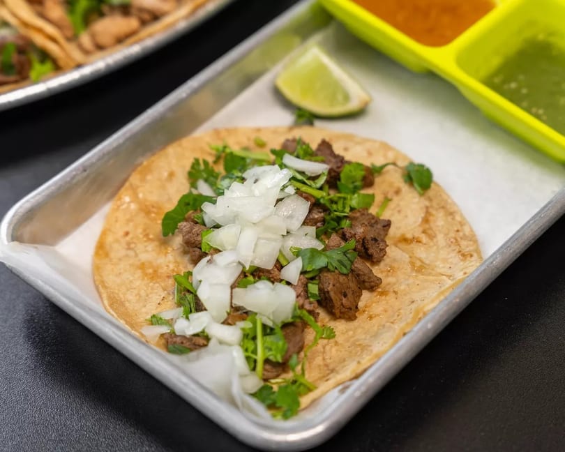 A single carne asada taco with onions and cilantro on a metal tray with lime and sauces.