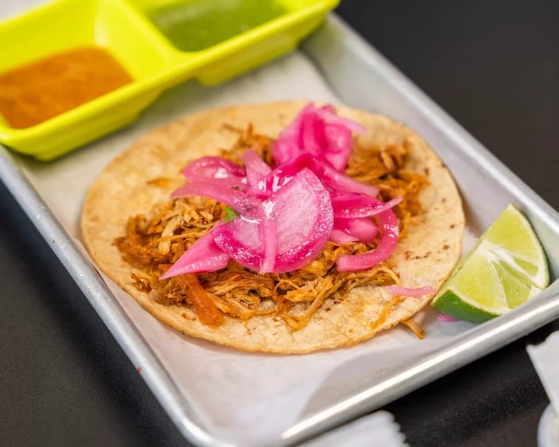 A single taco with shredded meat and pink pickled onions, served with lime and sauces.