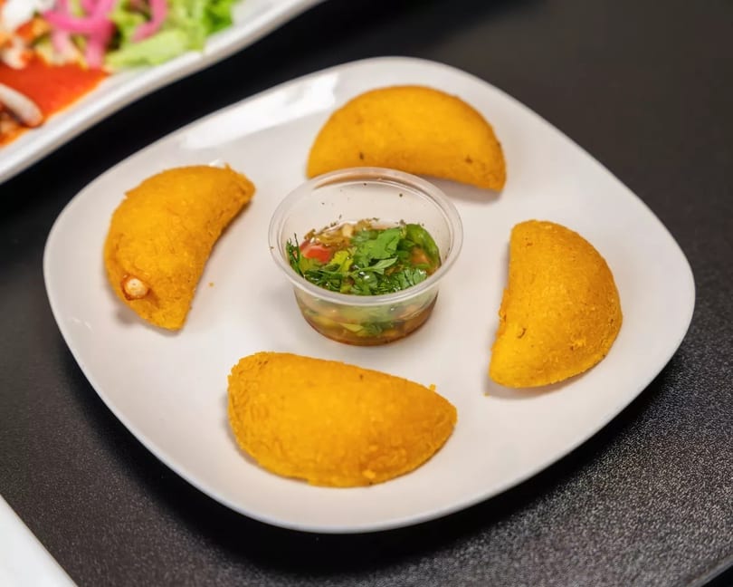 Four golden empanadas arranged around a small cup of green salsa on a white plate.