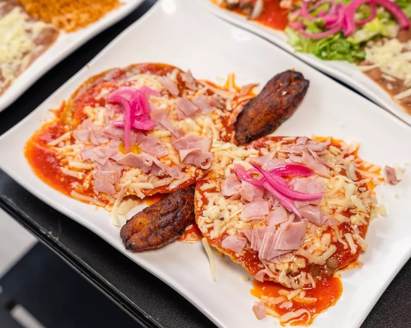 Two sopes topped with red sauce, cheese, ham, and pickled onions, with a fried plantain.