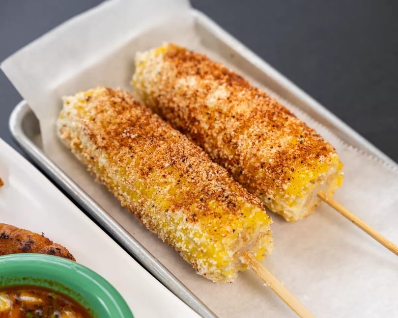 Two elotes, Mexican street corn on the cob, covered in cheese and chili powder on a tray.