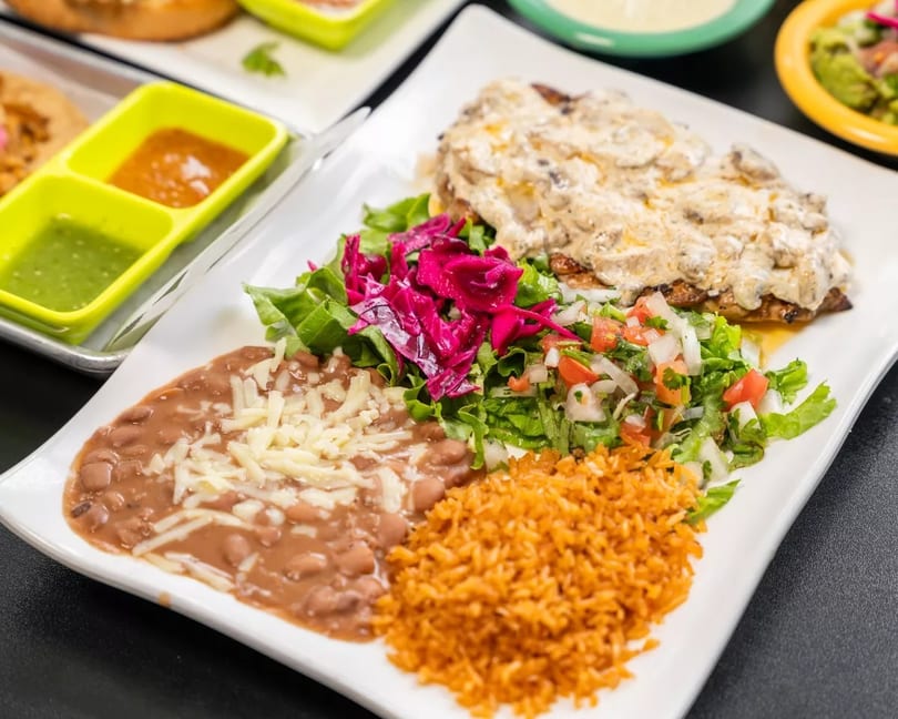 Chicken with mushroom sauce, rice, refried beans, and salad on a white plate.