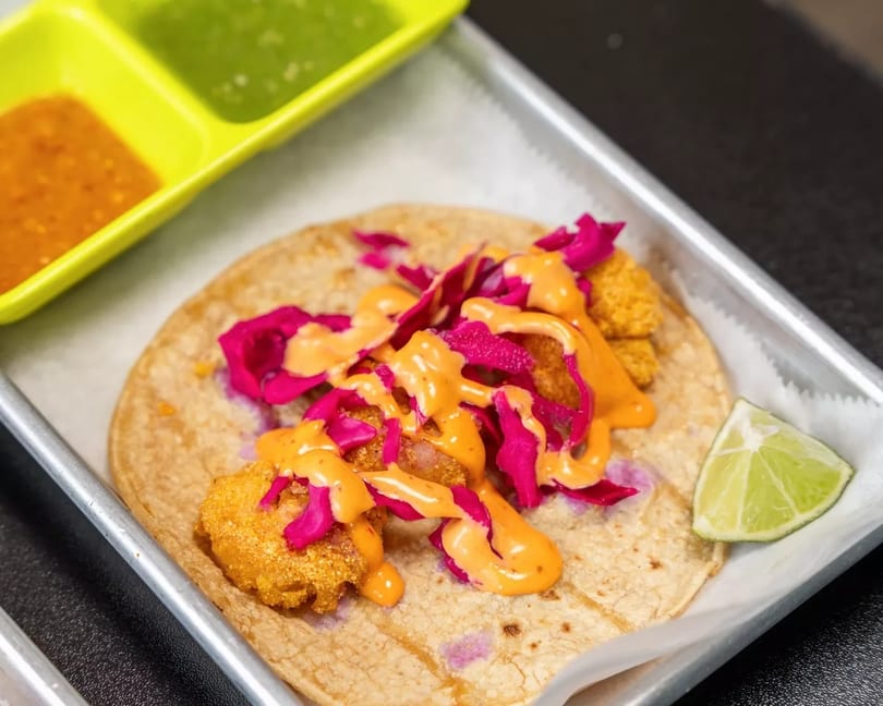 A shrimp taco with red cabbage and orange sauce, served with a lime wedge and two dipping sauces.
