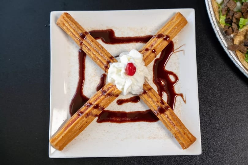 Four churros arranged in an X-shape on a white plate, with whipped cream and a cherry.